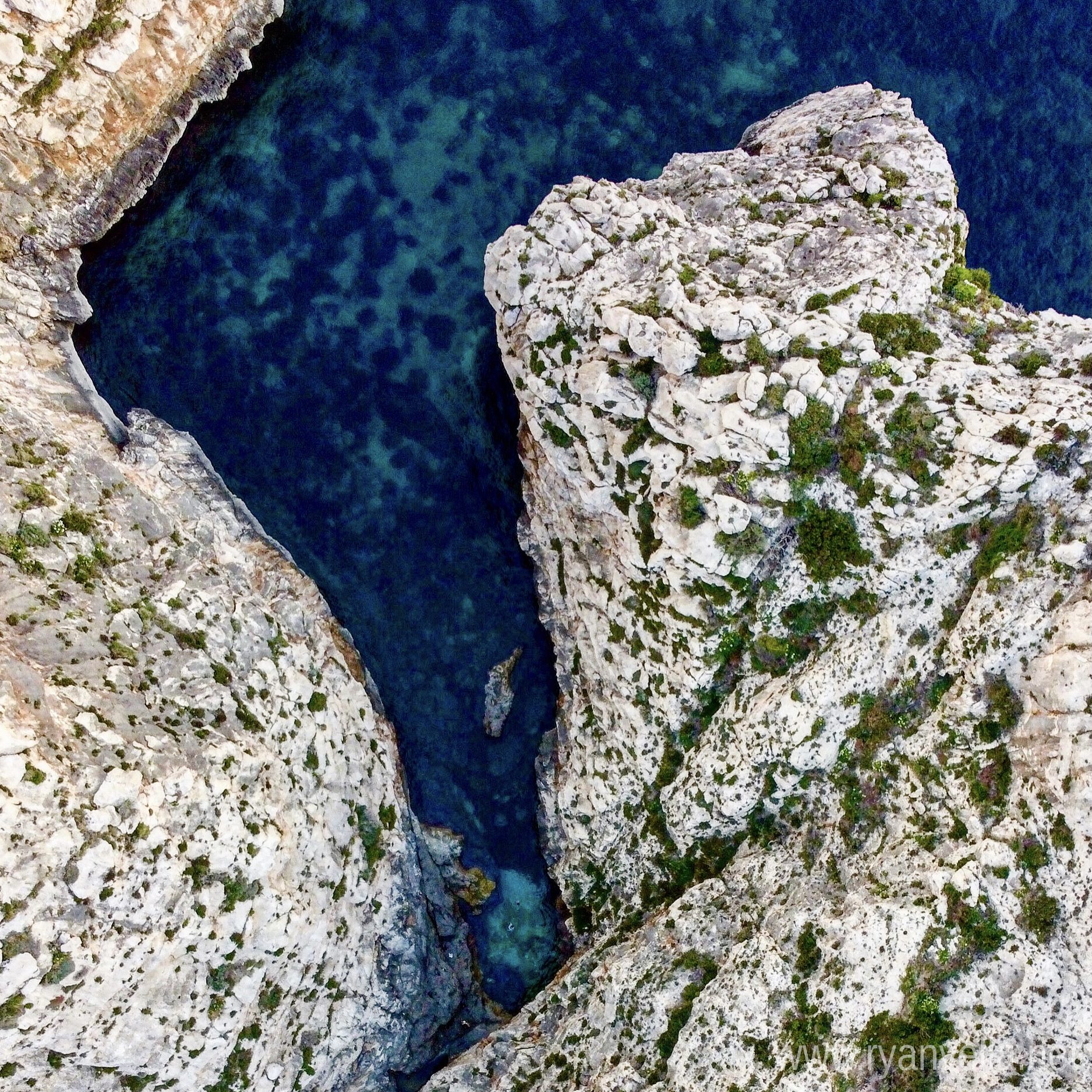 A narrow inlet of deep blue water between pale limestone cliffs, seen from directly above