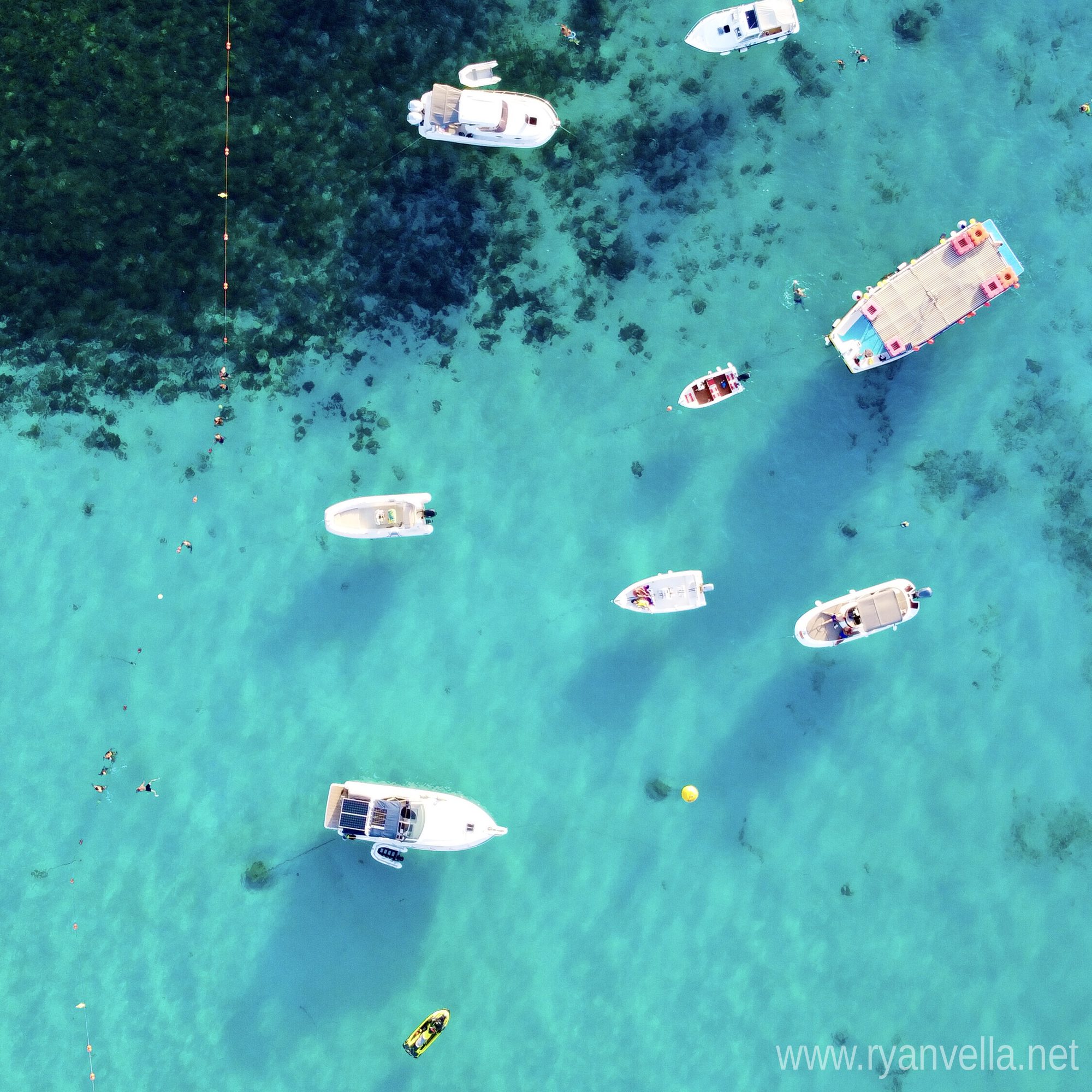 Small boats and a swimming pontoon moored over bright turquoise shallows, seen from directly above