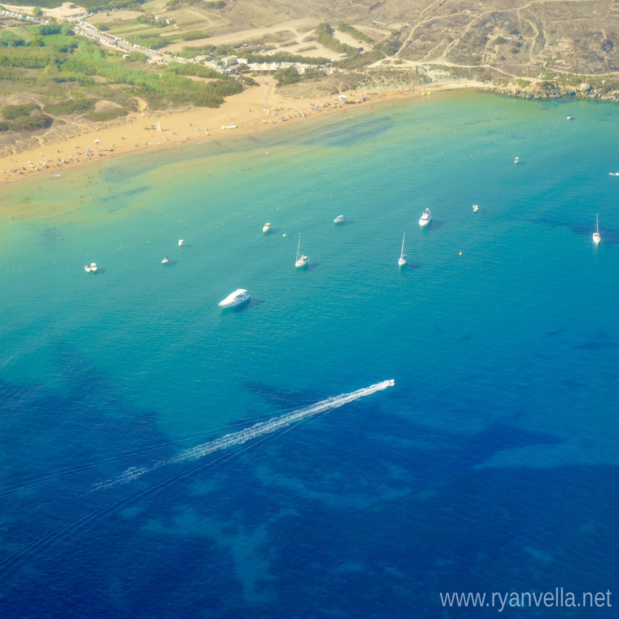 An aerial view of a sandy bay, boats anchored over turquoise shallows that deepen to open blue