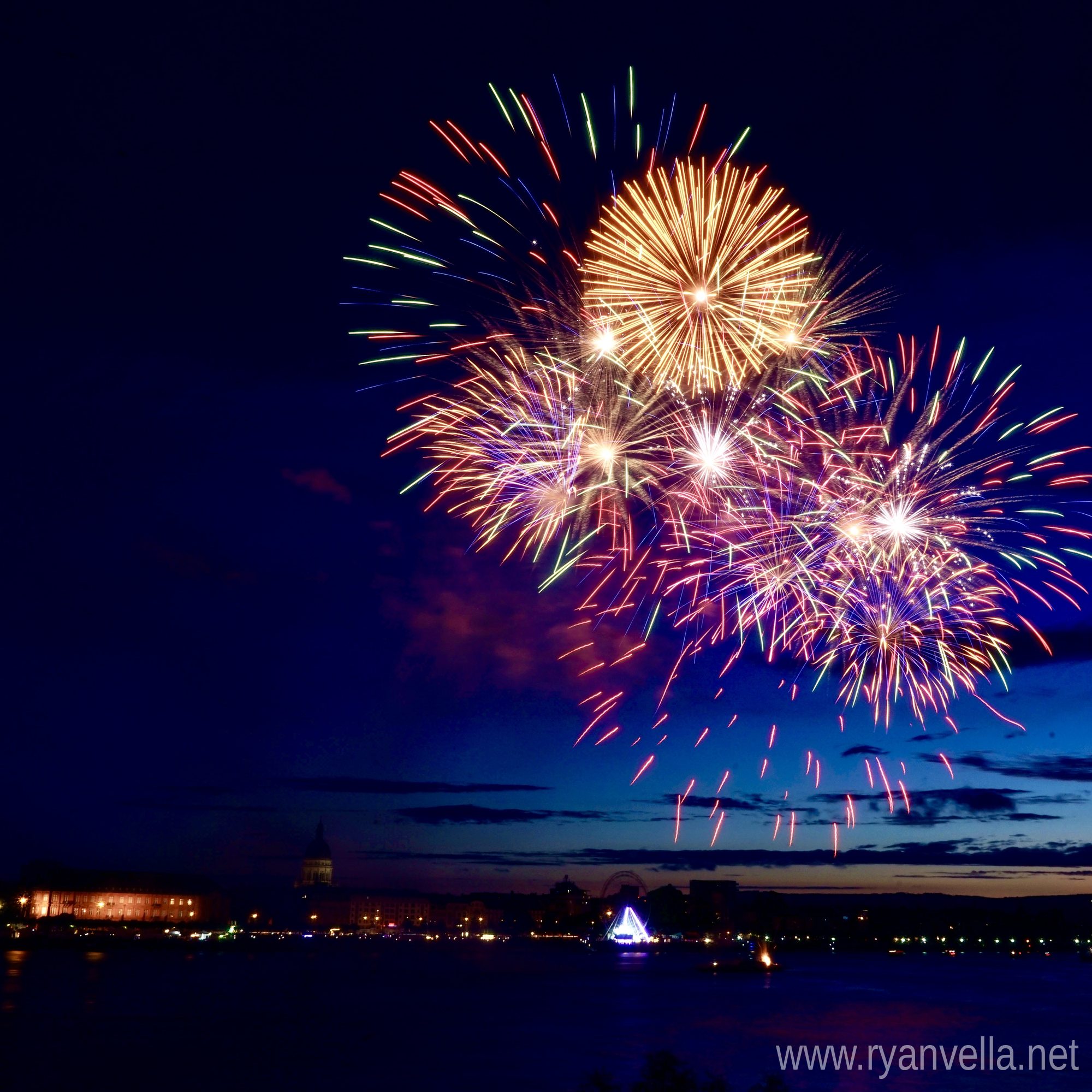Fireworks bursting in gold, red and green over a city waterfront at dusk